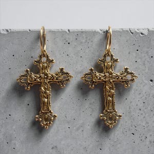 Puede incluir: Un par de pendientes de metal dorado con colgantes de cruz ornamentados. Cada cruz tiene detalles florales intrincados y bordes de filigrana, suspendidos de un aro circular simple. El metal tiene un acabado texturizado de estilo antiguo.