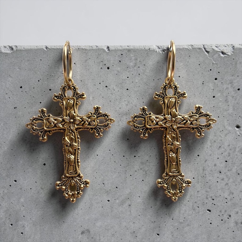 Puede incluir: Un par de pendientes de metal dorado con colgantes de cruz ornamentados. Cada cruz tiene detalles florales intrincados y bordes de filigrana, suspendidos de un aro circular simple. El metal tiene un acabado texturizado de estilo antiguo.