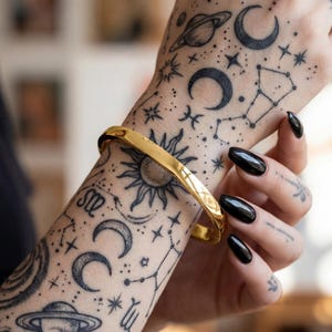 May include: A polished gold-tone metal bangle bracelet with a smooth, reflective surface and a simple, continuous circular band design. The bracelet is worn on a wrist that features extensive black ink celestial-themed tattoos of planets, moons, and stars.