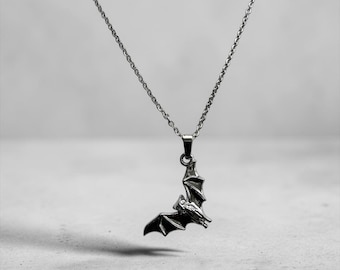 Può includere: Una collana in metallo color argento con un ciondolo a forma di pipistrello con le ali spiegate. Il ciondolo è sospeso a una sottile e delicata catenina color argento. Il design del pipistrello mostra strutture alari dettagliate e una piccola testa, realizzate con una finitura metallica lucida che riflette la luce.
