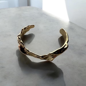 May include: A gold-colored cuff bracelet with an organic, textured design. The open-ended bracelet has an irregular, molten-like appearance, reflecting light and casting a shadow on the surface below. The jewelry piece is displayed on a light gray surface.