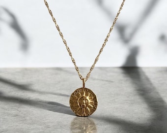 Può includere: Una collana color oro con una sottile catena ritorta e un ciondolo circolare. Il ciondolo presenta un design a rilievo con un volto solare, raggi radianti e intricate incisioni lungo il bordo esterno del medaglione a forma di moneta.