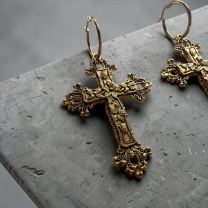 Puede incluir: Un par de pendientes de aro dorados con colgantes en forma de cruz ornamentados. Cada colgante presenta intrincados grabados florales y bordes de filigrana decorativos, suspendidos de un sencillo cierre de aro circular.
