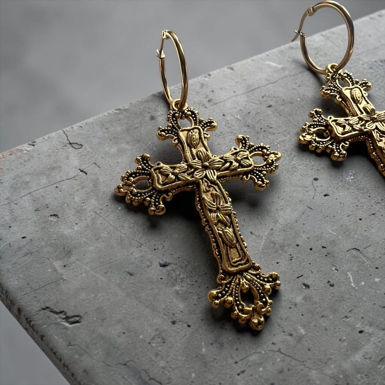 Puede incluir: Un par de pendientes de aro dorados con colgantes en forma de cruz ornamentados. Cada colgante presenta intrincados grabados florales y bordes de filigrana decorativos, suspendidos de un sencillo cierre de aro circular.