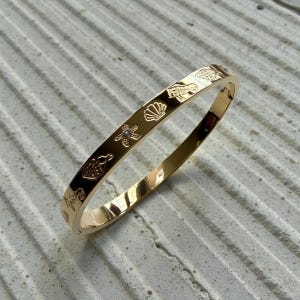 Może przedstawiać: Złota bransoletka z grawerowanymi wzorami o tematyce morskiej, w tym rozgwiazda, muszla i konik morski. Bransoletka ma gładkie, polerowane wykończenie i zawiasowe zapięcie. Bransoletka ma średnicę około 6,35 cm.