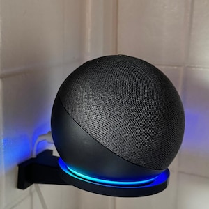 Pode incluir: Um alto-falante inteligente preto com exterior em malha de tecido, montado em um suporte de parede preto. O alto-falante tem um anel de luz azul na base. Um cabo de carregamento branco está conectado.