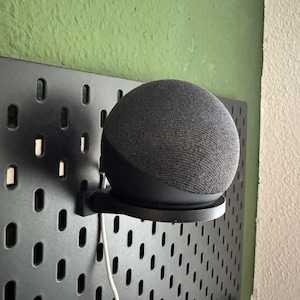 Peut inclure: Un haut-parleur intelligent noir monté sur un panneau perforé noir avec un cordon d'alimentation blanc. Le haut-parleur est une sphère recouverte de tissu gris foncé. Le panneau perforé est fixé à un mur vert.