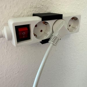 以下が含まれることがあります： 白の電源タップ。3つのコンセントと赤色のオン/オフスイッチがあります。黒いブラケットが白い壁にタップを固定しています。白い電源コードがコンセントに差し込まれています。