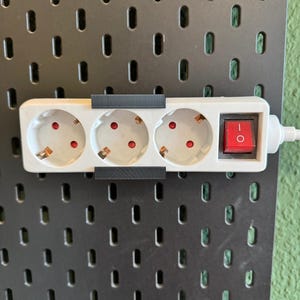Op de afbeelding: Een witte stekkerdoos met drie stopcontacten en een rode aan/uit-schakelaar. De stekkerdoos is gemonteerd op een zwart pegboard. Het pegboard heeft een reeks ovale gaten. De achtergrond is een groene muur.