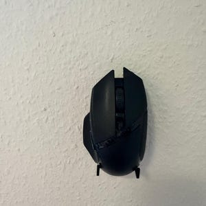 May include: A black wireless computer mouse with a scroll wheel and side buttons. The mouse is mounted on a white textured wall. The mouse has a sleek, ergonomic design.
