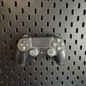 May include: A black PlayStation 4 controller with a directional pad, analog sticks, and action buttons. The controller has a touchpad and buttons labeled "Share" and "Options". It is set against a black pegboard background.