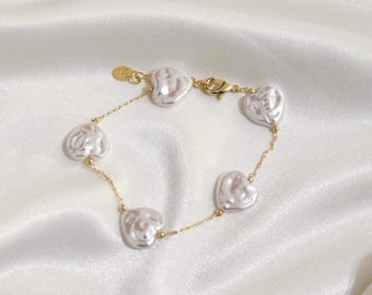 May include: A gold-toned bracelet featuring five heart-shaped, white pearls. The pearls are connected by a delicate gold chain, with a small gold clasp and a circular tag. The bracelet rests on a white, satin-like surface.