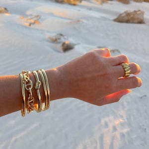 Puede incluir: Un conjunto de pulseras de oro apiladas en una muñeca, incluyendo una pulsera de cadena con un detalle de nudo. Un anillo de oro con un diseño en forma de corazón se lleva en el dedo anular.