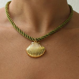 May include: A necklace featuring a gold-toned metal seashell pendant suspended from a thick, olive green twisted rope cord. The pendant has a ribbed texture and is attached to the cord with a small gold-toned metal jump ring.