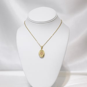 May include: A gold-colored oval locket necklace with a sunburst design. The pendant hangs from a delicate chain and is displayed on a white jewelry bust. The locket is a warm, metallic tone, and the chain is a matching gold color.