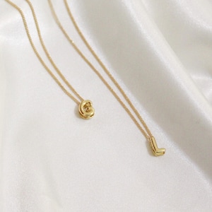 May include: Two gold-colored necklaces with delicate chains. One necklace features a pendant in the shape of the letter "G", and the other has a pendant shaped like the letter "L". The necklaces are displayed on a white, silky fabric.