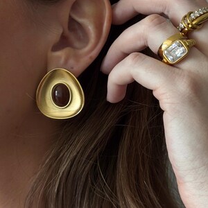 May include: Gold-toned earrings with a teardrop shape and a dark brown oval center. The earrings are worn on an ear, and several gold rings with clear stones are visible on a hand.