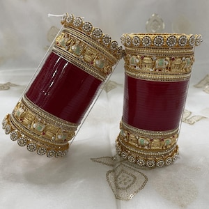 Può includere: Due braccialetti rossi ornati con abbellimenti color oro. I braccialetti presentano disegni intricati con piccoli accenti bianchi rotondi e motivi dettagliati. Le fasce dorate hanno piccole figure di elefanti e sono sormontate da una fila di accenti circolari.