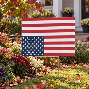 May include: A rectangular yard sign featuring a printed design of the United States flag with horizontal red and white stripes and a blue canton containing white stars. The sign is supported by two thin metal stakes inserted into the ground.