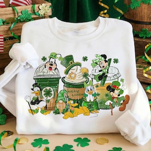 Puede incluir: Sudadera blanca con un diseño del Día de San Patricio con personajes de Disney. El gráfico incluye a Goofy, el Pato Donald, Minnie Mouse y Mickey Mouse, cada uno con detalles verdes y tréboles. También se ven las palabras "Lucky Charm".