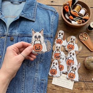 May include: A collection of embroidered iron-on patches featuring various dog and cat breeds dressed in white ghost costumes and holding orange jack-o'-lantern candy buckets. The patches are displayed alongside embroidery thread and scissors.