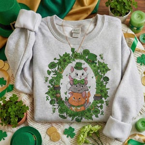 Puede incluir: Sudadera gris claro con un diseño del Día de San Patricio. El gráfico muestra tres gatos de dibujos animados con sombreros y lazos verdes, rodeados por una corona floral verde con tréboles y ollas de oro.