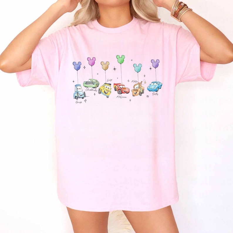 K&ouml;nnte beinhalten: Hellrosa T-Shirt mit einem bunten Cartoon-Autodesign. Jedes Auto hat eine andere Farbe und einen Namen darunter. Die Ballons &uuml;ber den Autos sind in Form von Mickey Mouse.
