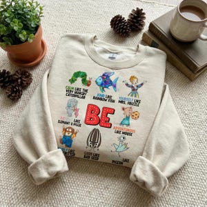 May include: Beige sweatshirt with colorful cartoon illustrations and the word "BE" in red. The graphics include a caterpillar, fish, Mrs. Frizzle, an elephant, a mouse, a critter, a seed, and a pigeon. The sweatshirt has long sleeves.