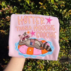 May include: Pink sweatshirt with the text "HOTTER THAN A HOOCHIE COOCHIE" in orange and pink. The graphic features a raccoon wearing heart-shaped sunglasses and a pearl necklace, lounging on a pink and orange inflatable ring, holding an American flag.