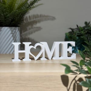 Freestanding 3D printed HOME sign with heart detail in white on a wooden shelf with green plants in the background