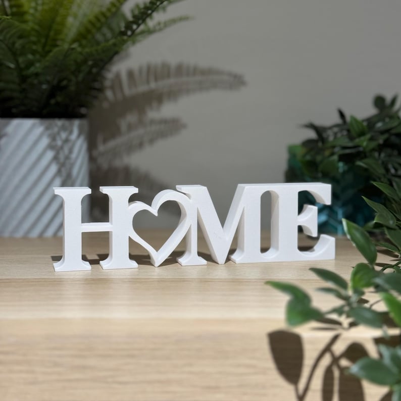 Freestanding 3D printed HOME sign with heart detail in white on a wooden shelf with green plants in the background