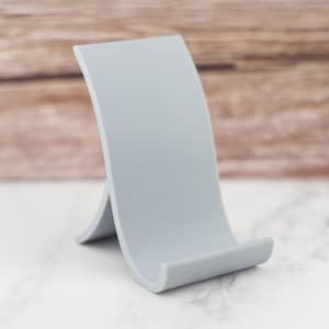 May include: A grey phone stand made of plastic. The stand has a curved design and a wide base.