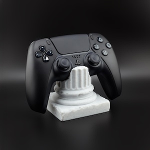 May include: A black wireless games controller sits on a white marble-effect stand. The controller features various buttons and joysticks. The stand is a miniature replica of a classical column.