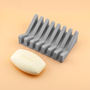 May include: A grey soap dish with a slotted design, alongside a bar of soap. The soap dish is made of a durable material and is designed to allow water to drain, extending the life of the soap. The bar of soap has the word "Vaseline" printed on it.