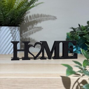 3D printed HOME sign with heart detail in black standing on a light wooden surface with plants in the background