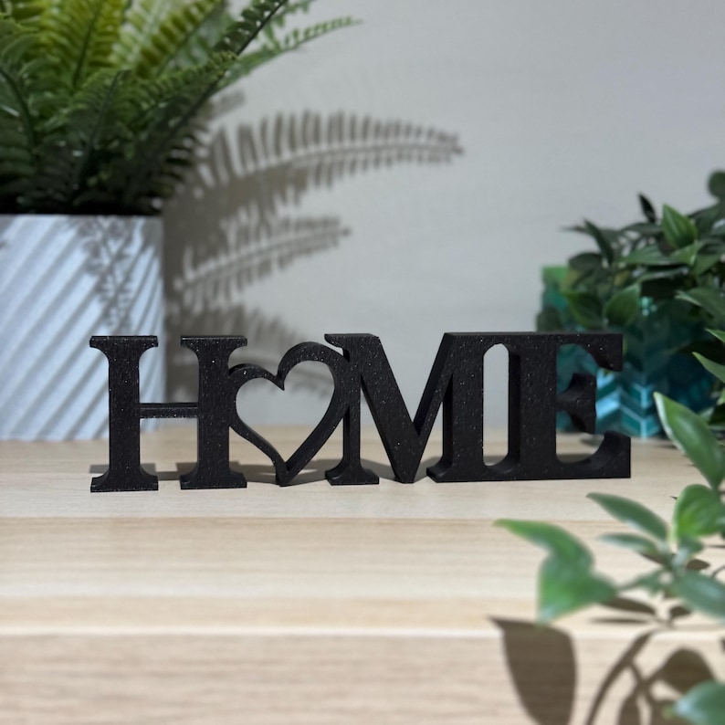 3D printed HOME sign with heart detail in black standing on a light wooden surface with plants in the background