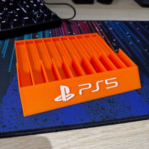 May include: Orange plastic game case with slots for storing video game discs. The case has the PlayStation logo and the text "PS5" printed on the front.