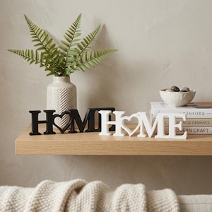 Freestanding 3D printed HOME signs with heart detail in black and white displayed side by side on a oak shelf with a white textured vase of fern leaves, stacked books and a small stone bowl in the background