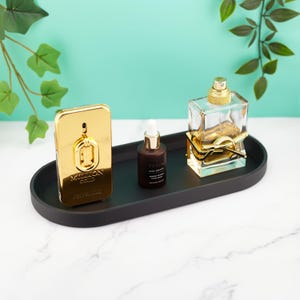 May include: A gold perfume bottle with "Million Gold" text and a gold pendant. A small brown bottle with a white dropper and "Tropic Sun Drops" text. A clear perfume bottle with a gold cap and accents. All items are on a black oval tray.