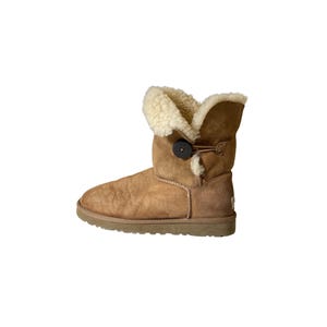 May include: A brown suede boot with a cream-colored shearling lining and a button closure. The boot has a worn appearance and a rubber sole. The boot is designed for cold weather and casual wear.
