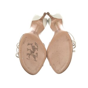 May include: A pair of white high-heeled sandals with thin silver straps. The heels are a light brown colour. The soles of the shoes are a light tan colour. The shoes are set against a white background.