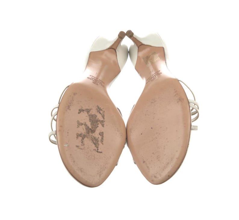 May include: A pair of white high-heeled sandals with thin silver straps. The heels are a light brown colour. The soles of the shoes are a light tan colour. The shoes are set against a white background.