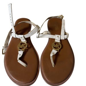 May include: Pair of white and gold Michael Kors sandals. The sandals have a brown footbed, white straps, and a gold MK logo. The sandals are designed with a thong style and adjustable ankle straps.
