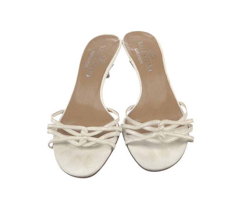 May include: A pair of off-white Valentino Garavani heeled sandals. The sandals have thin straps across the toes and a small heel. The Valentino Garavani logo is visible on the insole. The shoes are set against a white background.