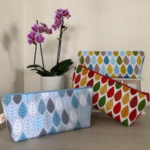 May include: Three zippered pouches with a leaf pattern. The pouches are in shades of blue, red and yellow. The pouches are lined up on a wooden surface.