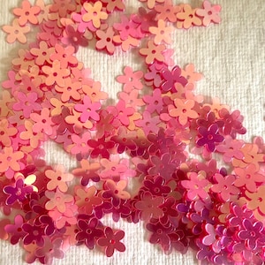 May include: A large quantity of pink iridescent flower-shaped sequins. The sequins are scattered on a white fabric surface.