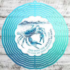 May include: A blue crab wind spinner with a scalloped edge and a circular design. The crab is depicted in shades of blue with the text "AND INTO THE OCEAN I GO TO LOSE MY MIND AND FIND MY SOUL."