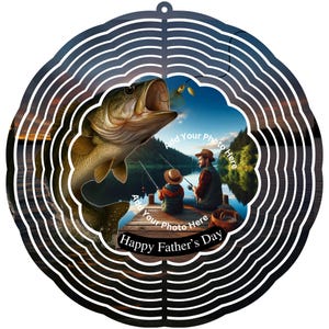 May include: A circular metal wind spinner with a fishing theme. The design shows a large fish leaping towards a lure, with a father and child fishing on a dock. Text reads "Happy Father's Day".