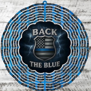May include: A round, blue and silver metal wind spinner with the text "BACK THE BLUE". The design features a police badge with the American flag and a blue line, set against a dark blue background with lightning.