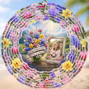 Op de afbeelding: Een decoratieve windspinner met een bloemenontwerp en een centraal beeld van een vrouw die bloemen vasthoudt. De spinner heeft een gelaagd ontwerp met roze, gele, paarse en blauwe bloemen. De tekst "Happy Mother's Day Up In Heaven" is zichtbaar.